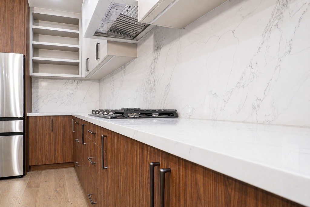White marble-look backsplash and range hood above a cooktop, wood cabinets and pale counter