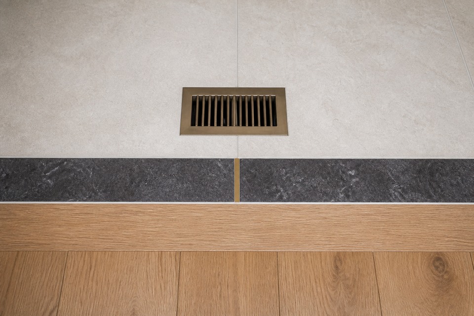 Close view of a floor register aligned with pale tile, a dark border and oak